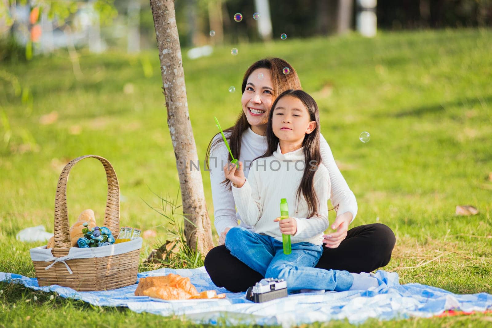 Happy Asian mother and little girl daughter child having fun and enjoying outdoor together sitting on the grass blowing soap bubbles during a picnic in the garden park on a sunny day family time