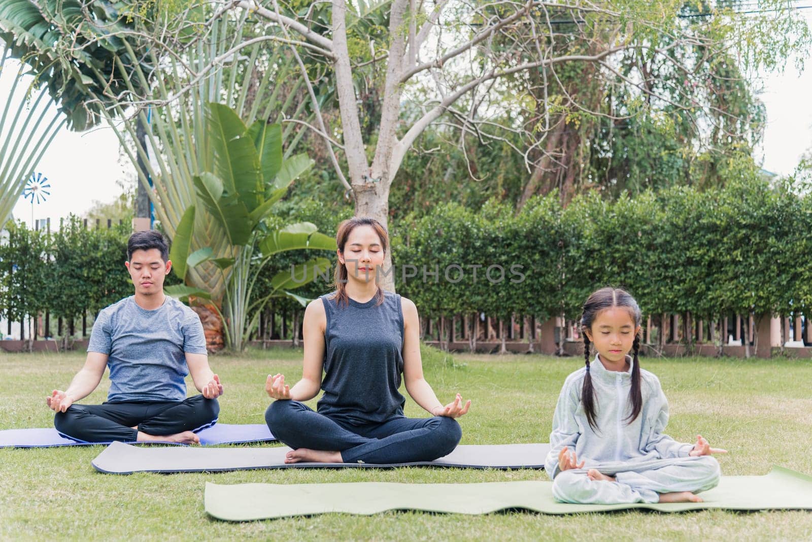 Asian young mother, father practicing doing yoga exercises with child daughter outdoors in meditate pose together in nature a field garden park, family kid sport and exercises for healthy lifestyle