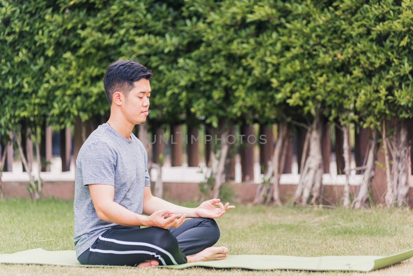 Asian young man doing yoga outdoors in meditate lotus pose sitting on green grass with closed eyes at the garden park, health care concept