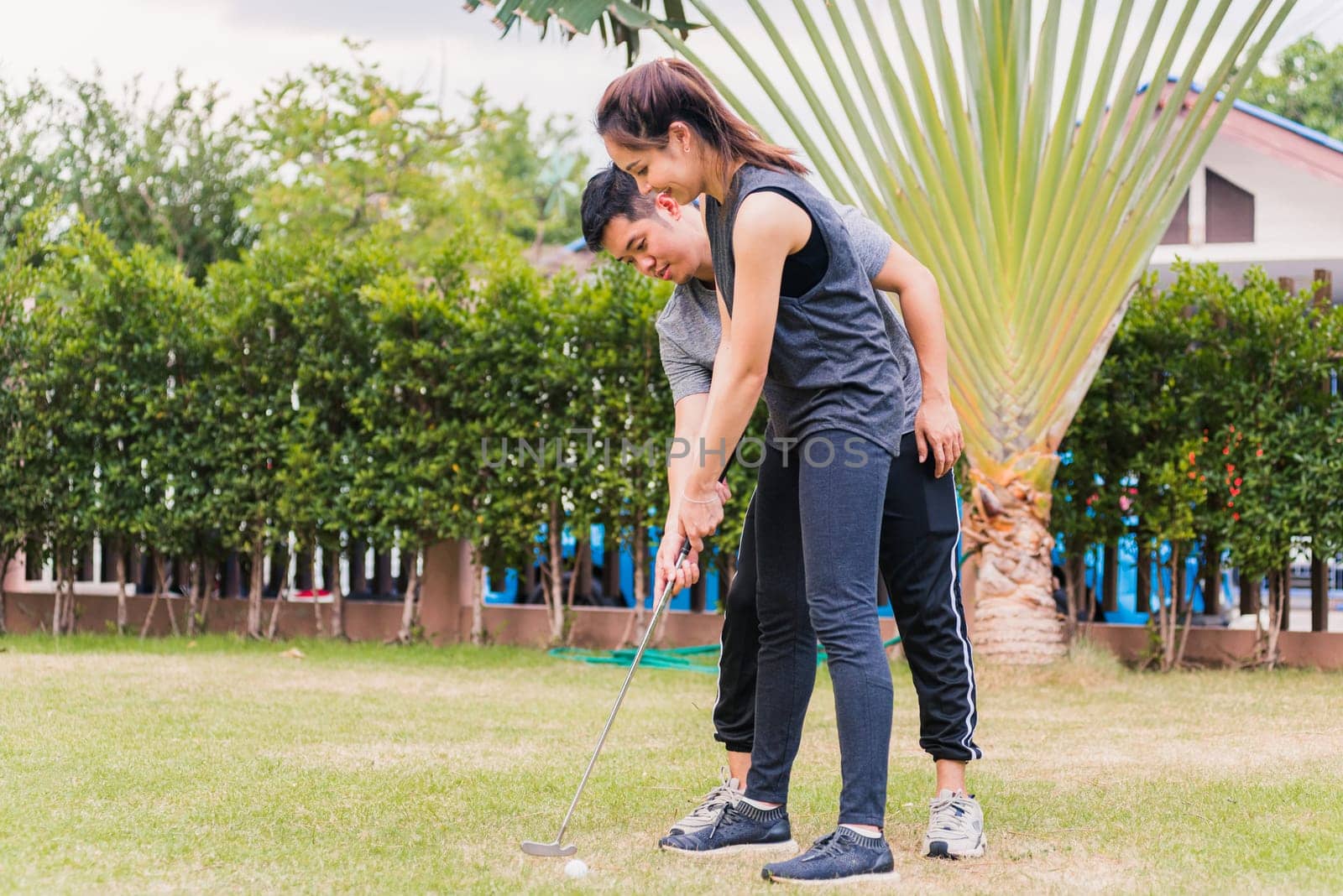 Asian young man support teaching training woman to play perfect golf while standing together in nature a field garden park. Couple trainer giving a lesson on the golf course
