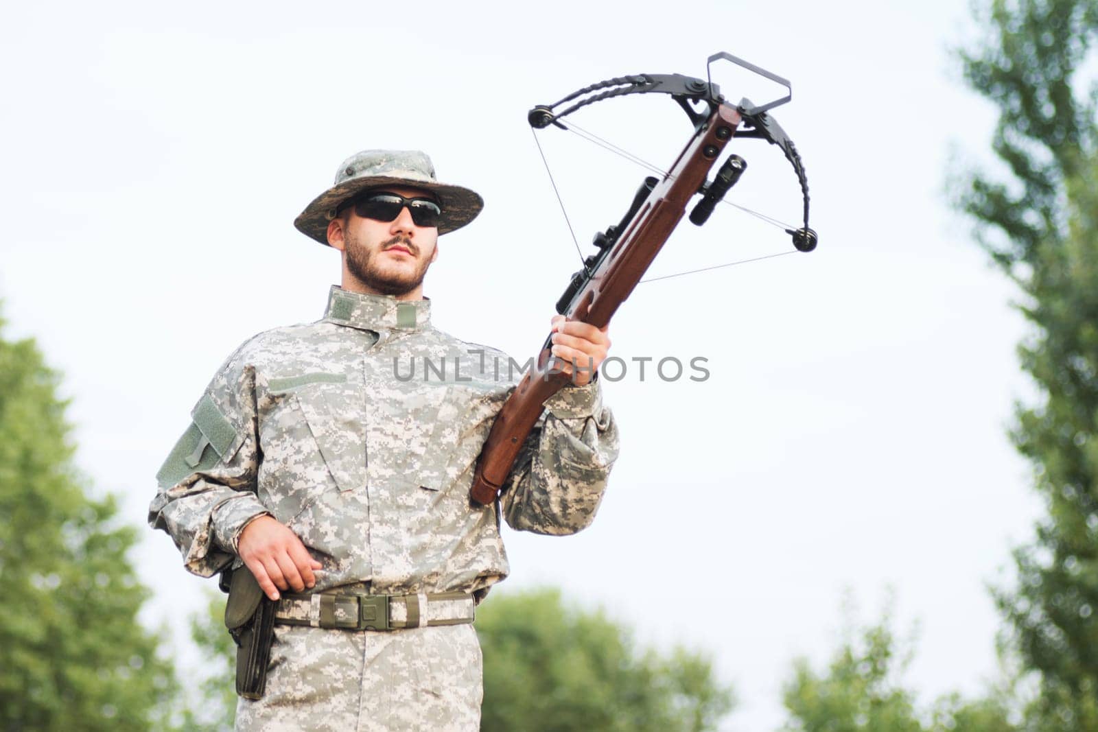 The hunter or army soldier in military uniform is holding a crossbow weapon outdoors.