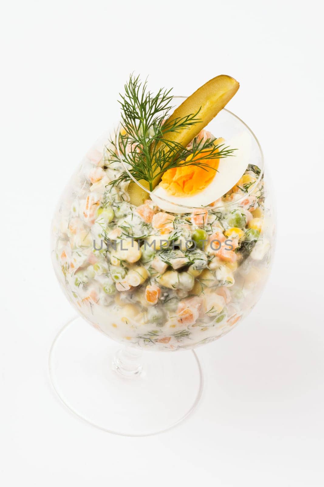 Russian salad with garnish is served in drinking glass on white background.
