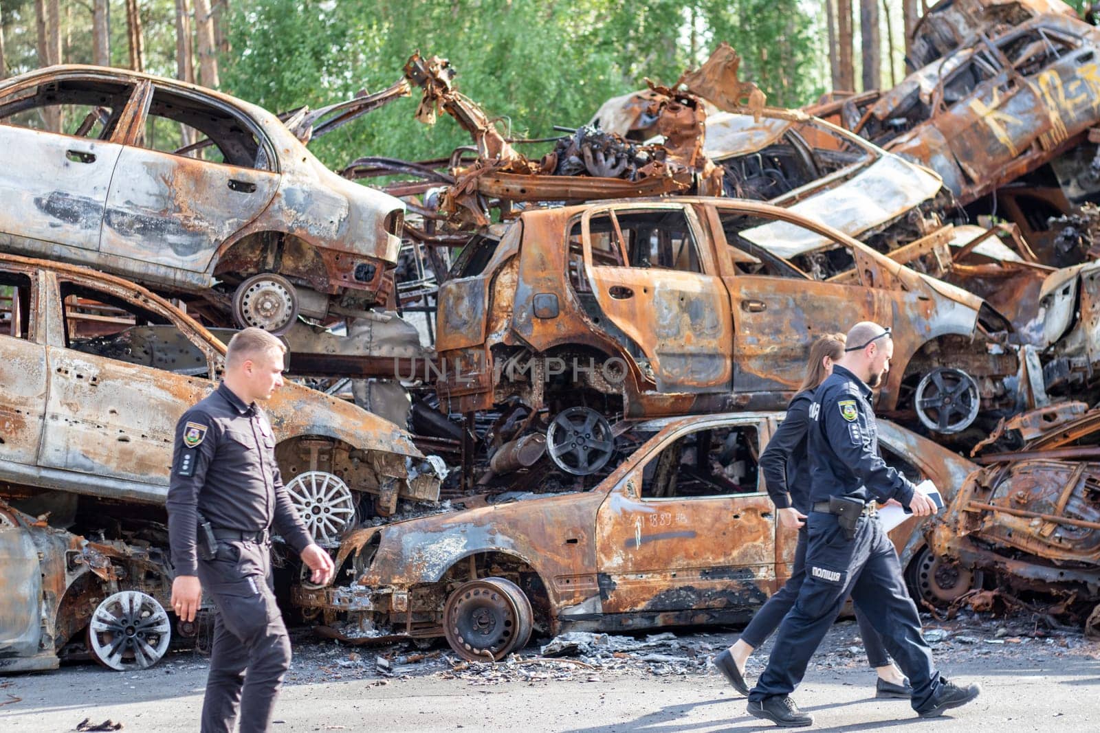 Police officers in front of burnt cars destroyed by rocket explosions. War in Ukraine. Destroyed vehicles of civilians. Car graveyard. Ukraine, Irpen - May 12, 2022