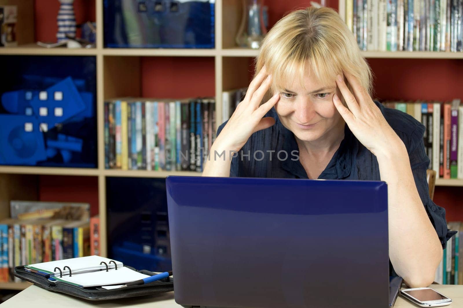 The middle aged woman is sitting at home and is working with a notebook. She is smiling and surprised by what she reads in the notebook. All potential trademarks are removed.