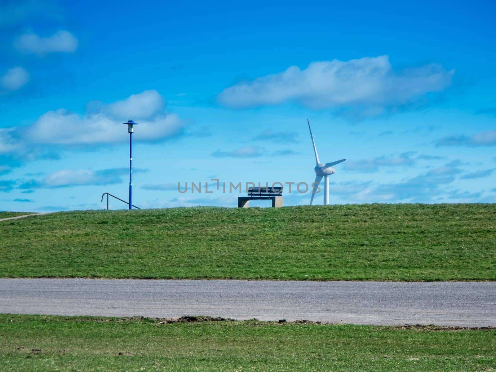 Landscape view of the German North Sea coast with a park bench on the dyke and a street lamp next to it, as well as a wind turbine in the background against a blue sky with a few clouds.