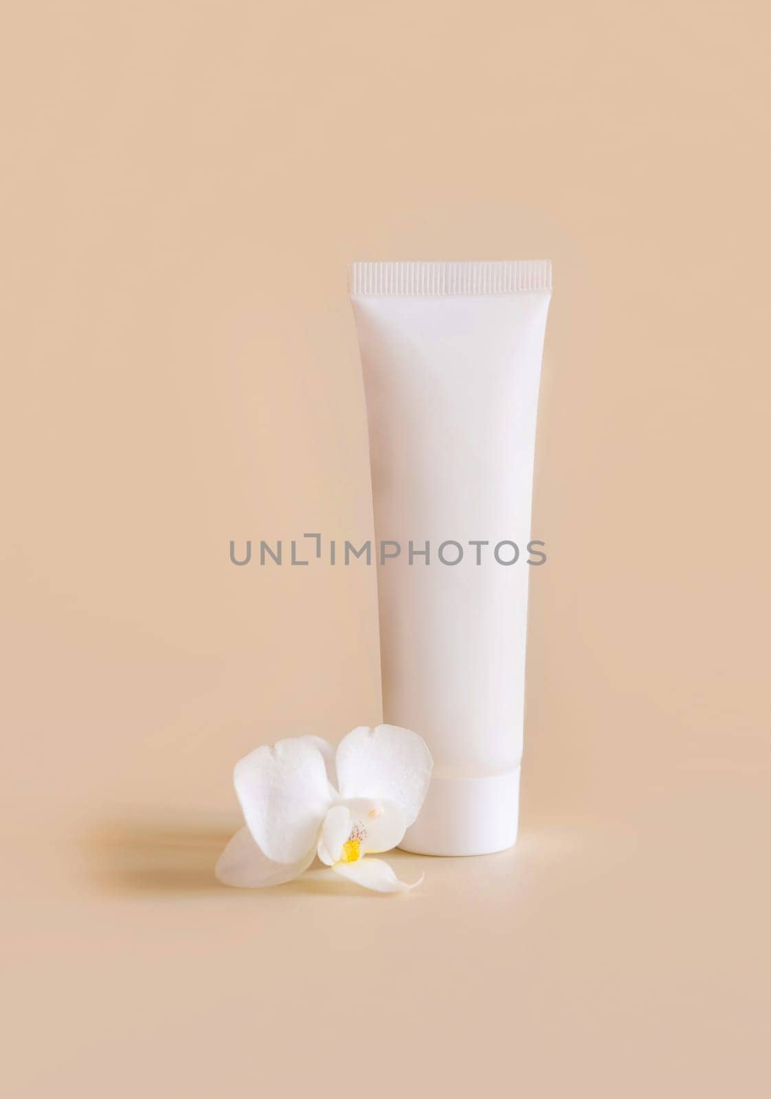 White plastic cream tube near white orchid flower on light yellow, close up, mockup. Skincare beauty product, cream or lotion. Exotic natural cosmetics, pastel minimal composition..