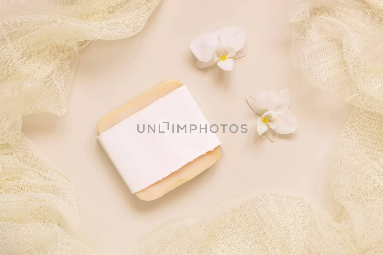 Hanmade soap bar near white orchid flowers and tulle on light yellow, top view, mockup. Skincare beauty product. Exotic natural cosmetics, pastel minimal composition..