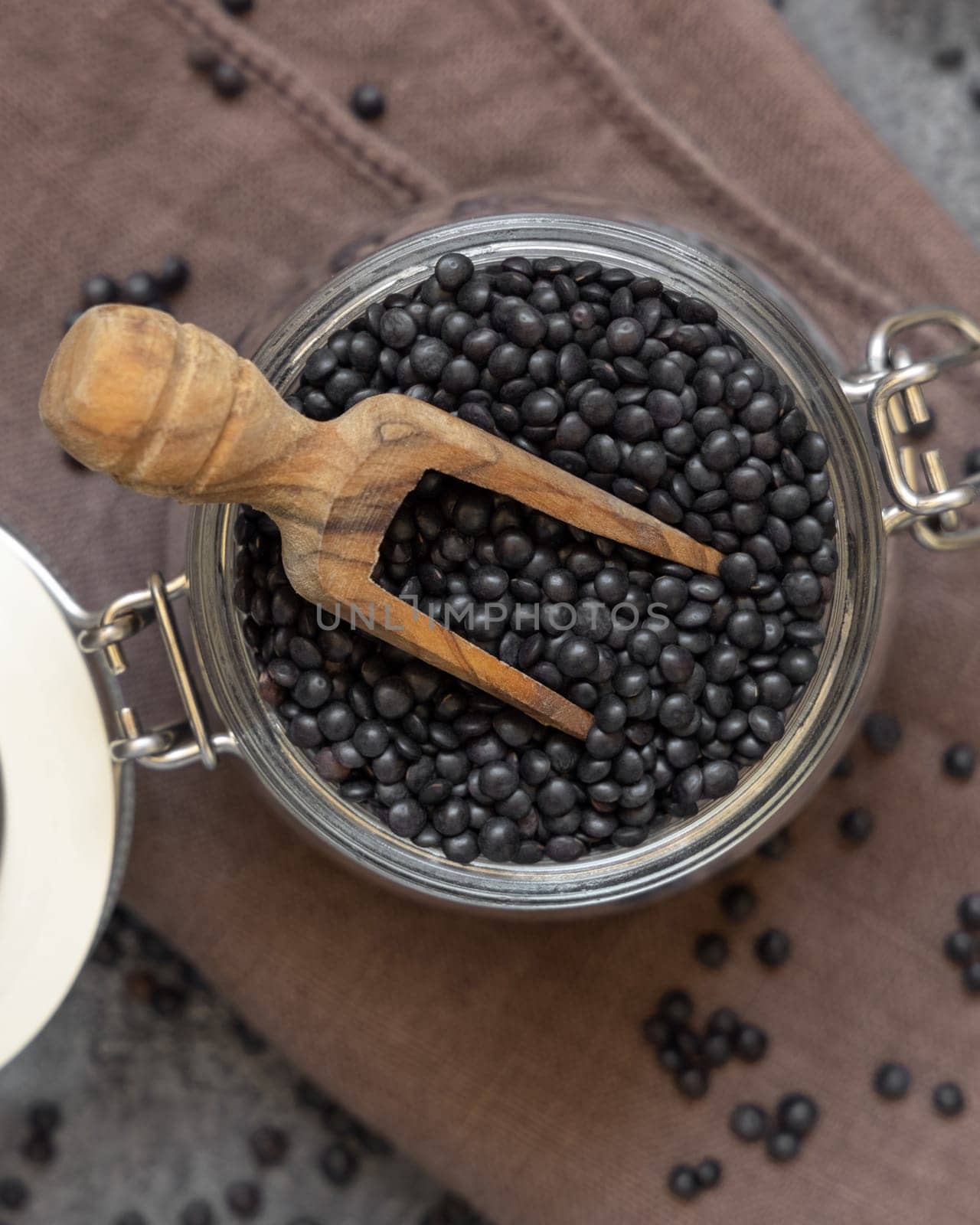 Glass jar of dry black lentils beans with a wooden scoop on grey table top view. Healthy eating, protein source for vegan and vegetarian diet. Traditional Asian and North African cuisines ingredient