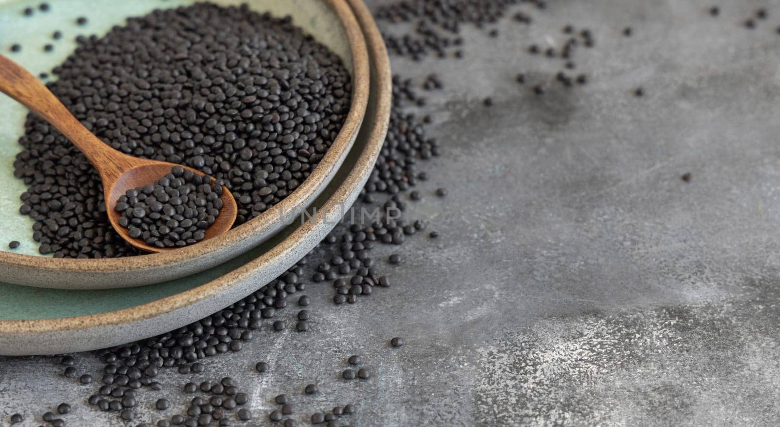 Plate of dry black lentils beans with a spoon on grey table close up, copy space. Healthy eating, protein source for vegan and vegetarian diet. Traditional Asian and North African cuisines ingredient