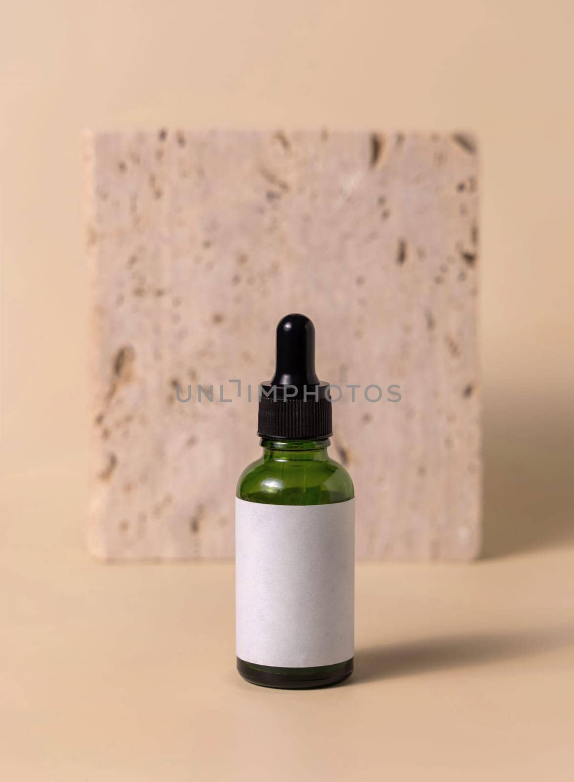 Green glass dropper bottle near beige travertine stone on light yellow, close up, mockup. Skincare beauty product, serum or lotion. Handmade natural cosmetics, trendy minimal composition..