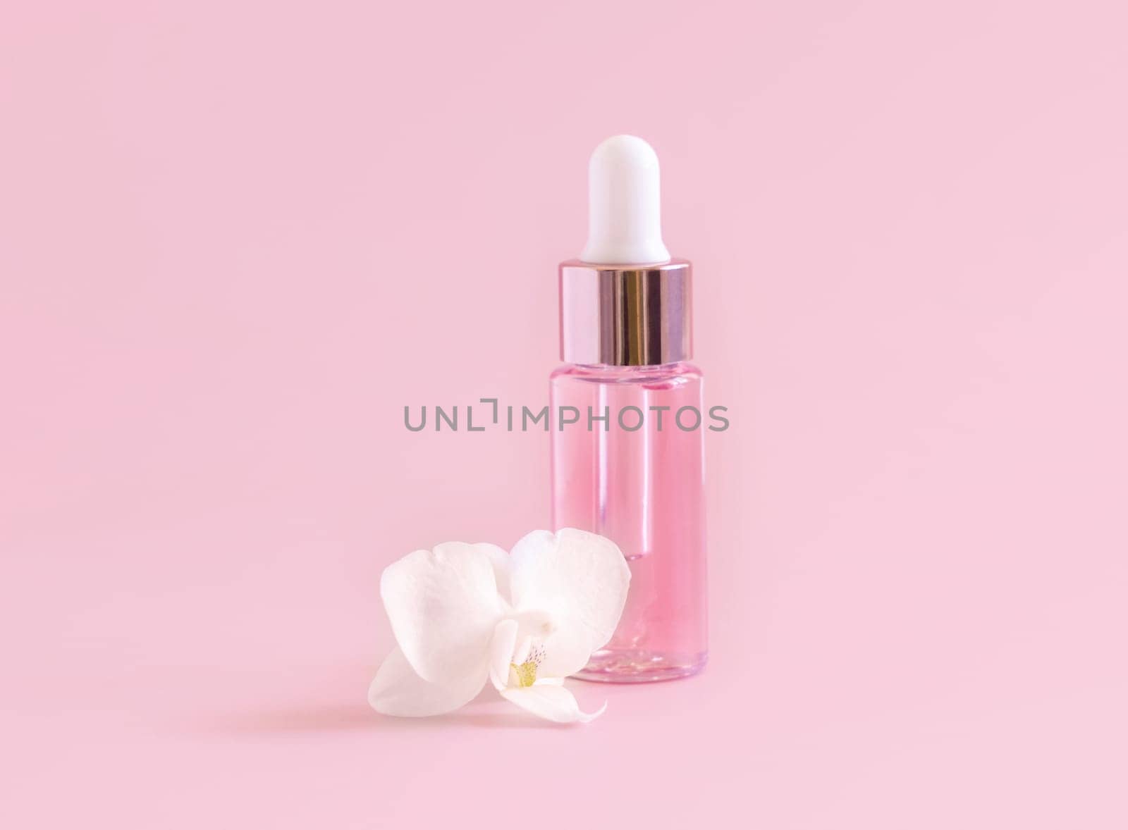 Light Pink Dropper Bottle near white orchid flower on light pink ,close up, mockup. Skincare beauty product, Aromatic oil or serum. Exotic cosmetics, pastel minimal composition..