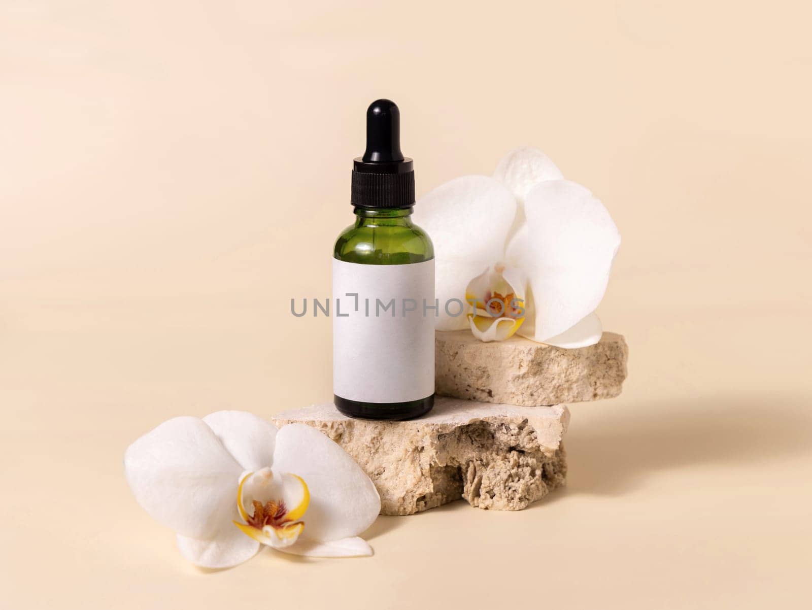 Green glass dropper bottle on stone near white orchid flowers on light yellow, close up, mockup. Skincare handmade beauty product, serum or lotion. Exotic natural cosmetics..