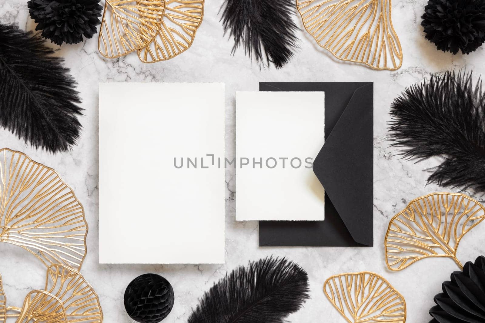 Blank cards and black envelope on marble table near black feathers and golden leaves top view. Wedding set mockup. Elegant femminine suite with paper cards, flat lay