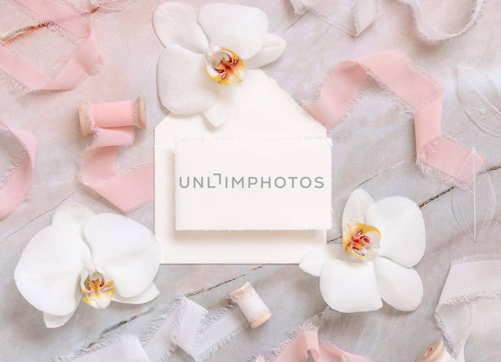 Wedding horizontal card and envelope near white orchid flowers and silk ribbons on marble top view, mockup. Romantic scene flat lay with handmade paper card