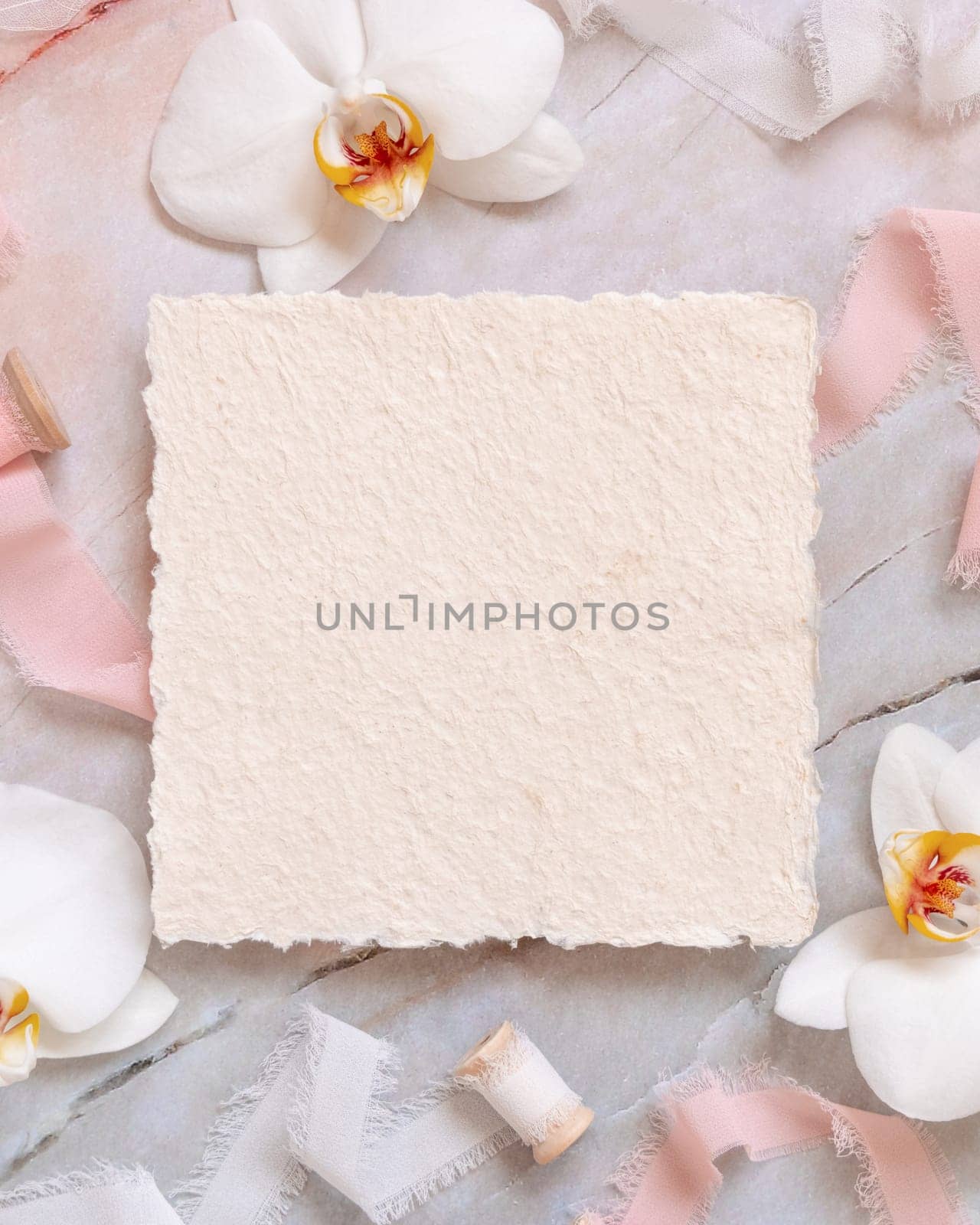 Wedding square card near white orchid flowers and silk ribbons on marble top view, mockup. Romantic scene flat lay with handmade paper card. Valentines or Spring romantic concept