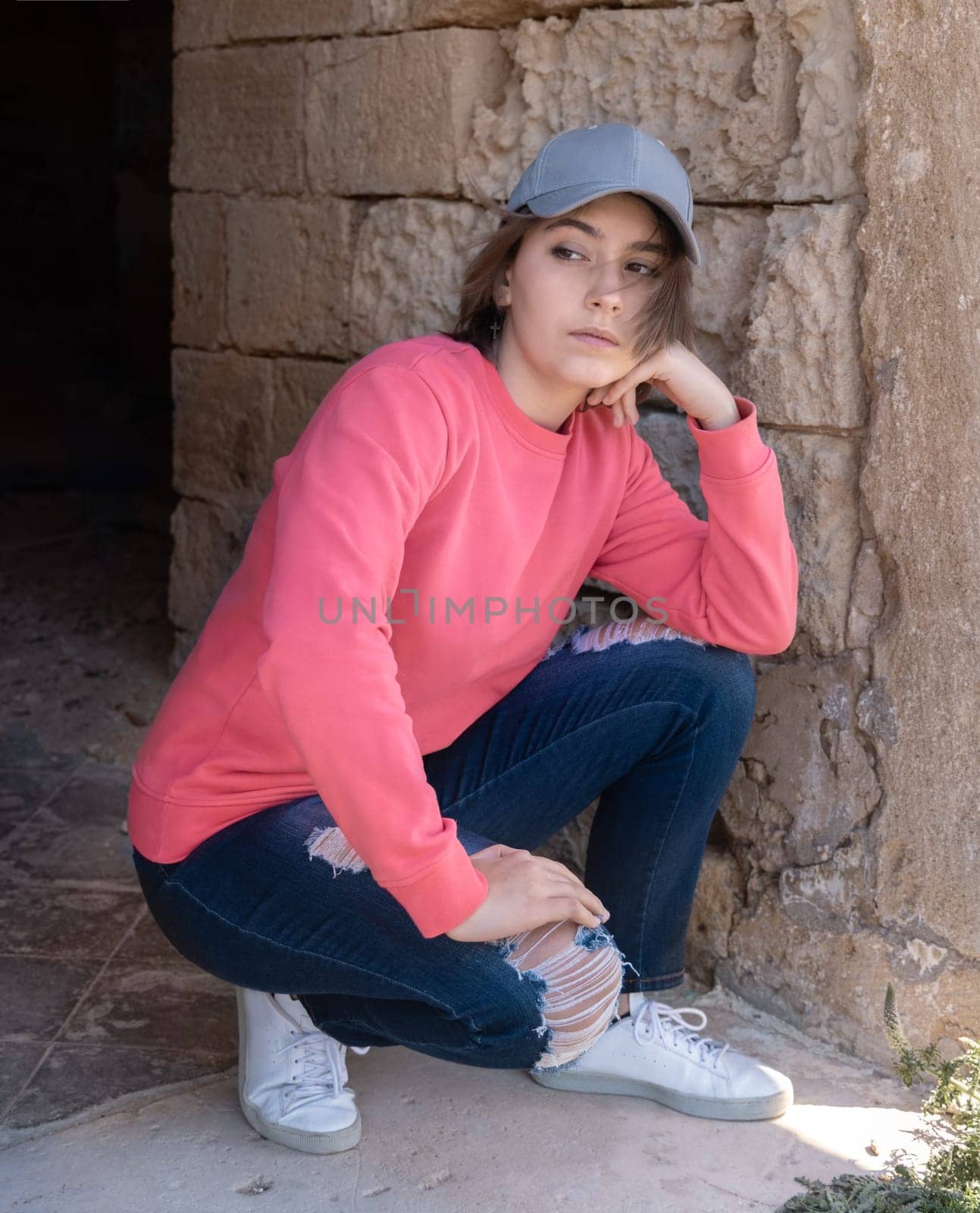 Teen girl wearing pink long sleeve shirt and baseball cap looking aside. Mockup. Serious Teenager girl seating on her haunches outdoor, spending her time alone. Adolescence concept