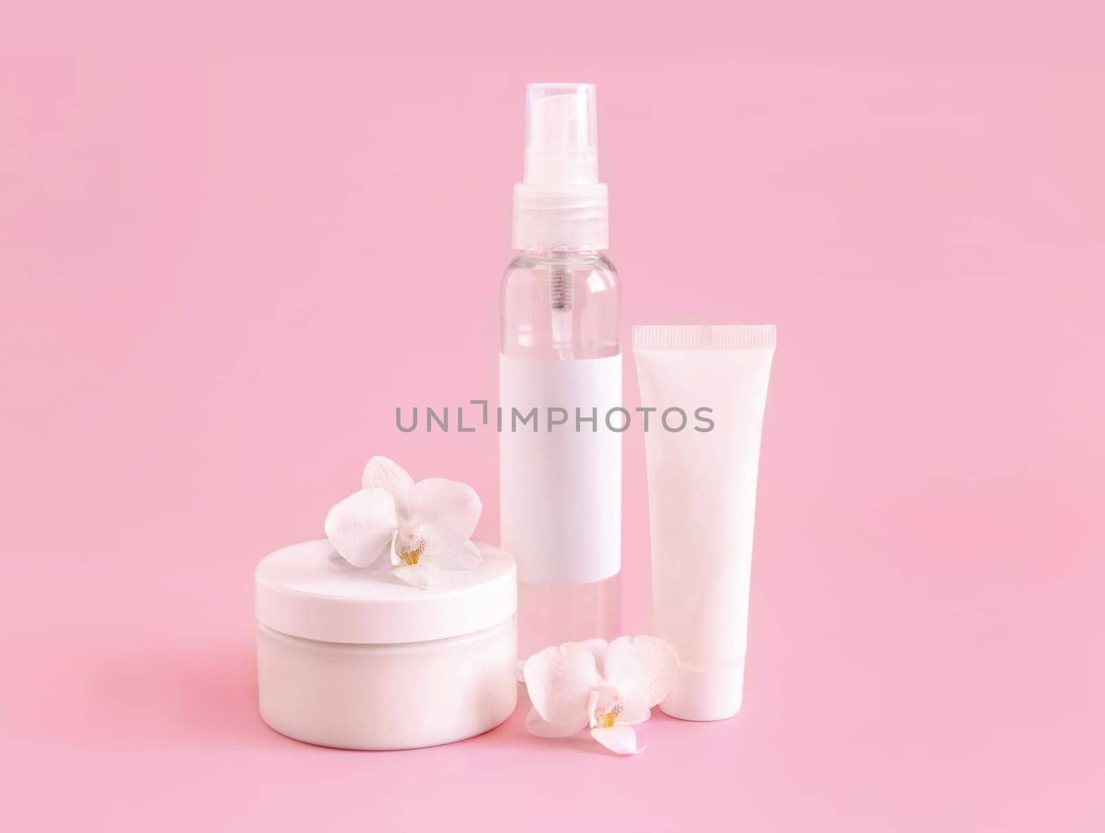 White cosmetic jar, tube and bottle near white orchid flower on light pink, close up, mockup. Skincare beauty product, cream or lotion. Exotic natural cosmetics, pastel minimal composition..