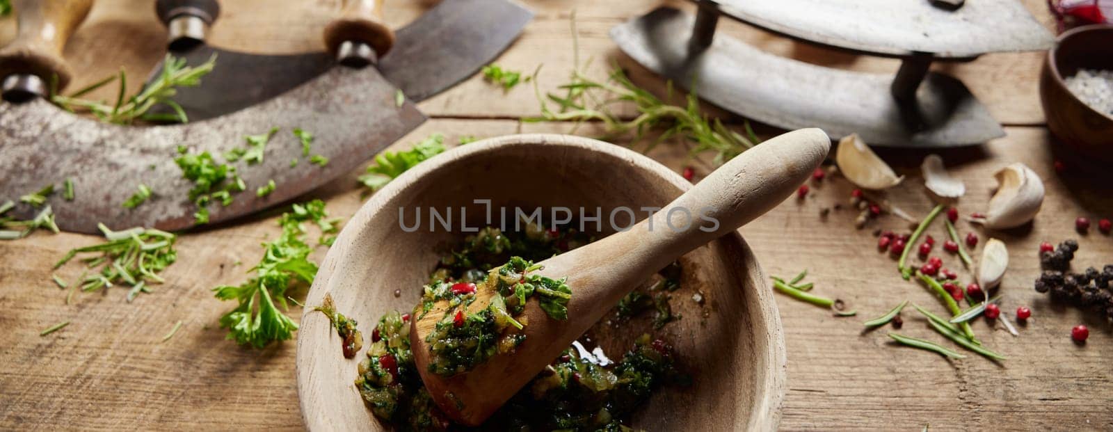 From above of wooden mortar with grinded greens placed on messy table with old fashioned sharp knives in light kitchen