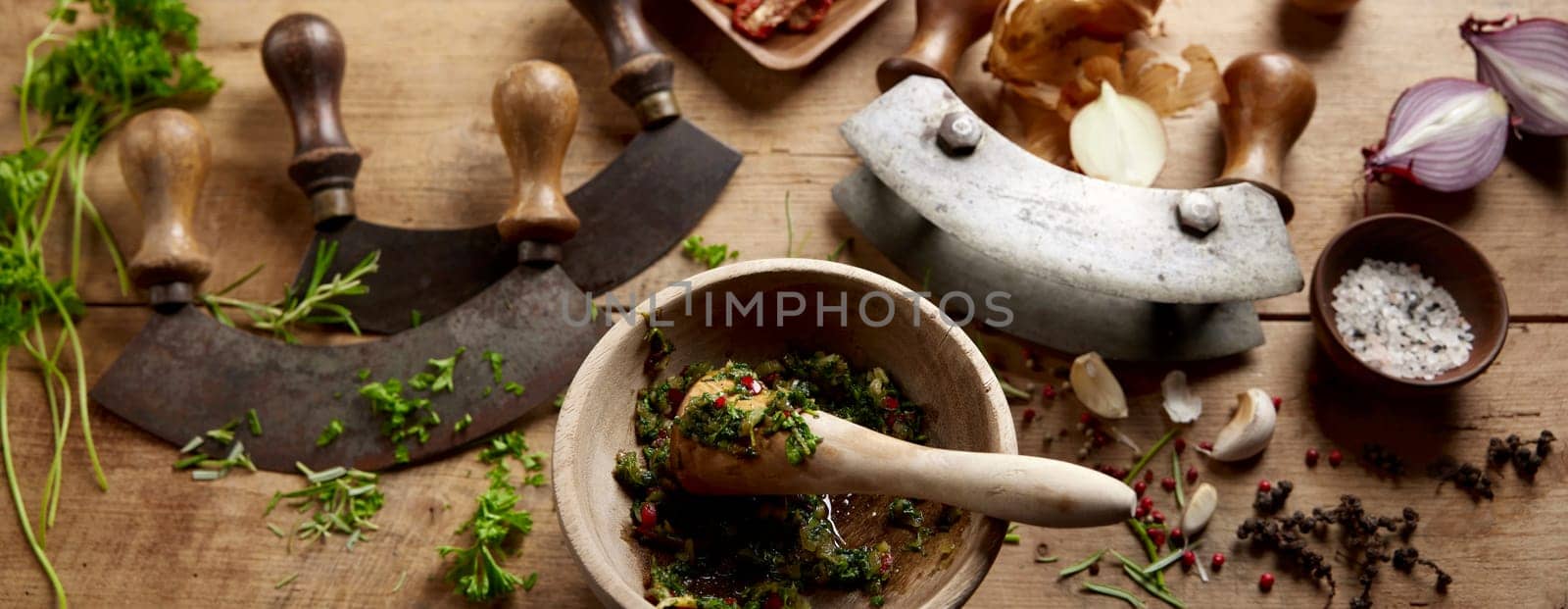 From above of mortar and pestle with grinded herbs placed on wooden table with old fashioned mezzaluna knives in kitchen