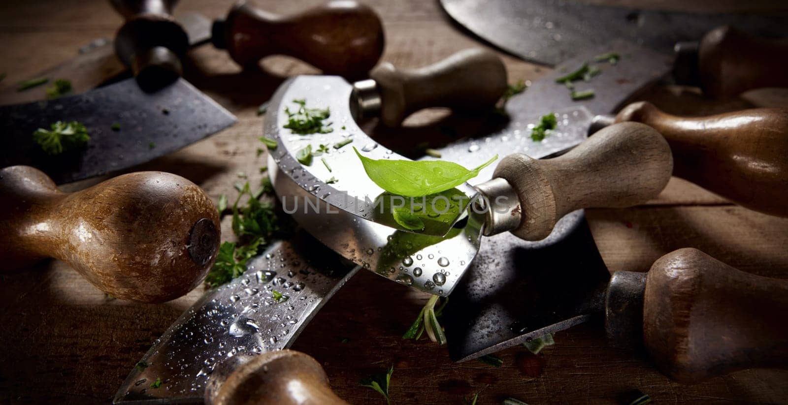 Collection of old fashioned mezzaluna knives with wooden handles and curved blades covered with herbs placed on table in kitchen