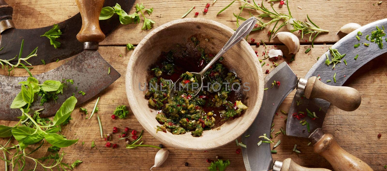 From above of mortar with freshly grinded herbs placed on wooden table with old fashioned traditional chopping knives in light kitchen