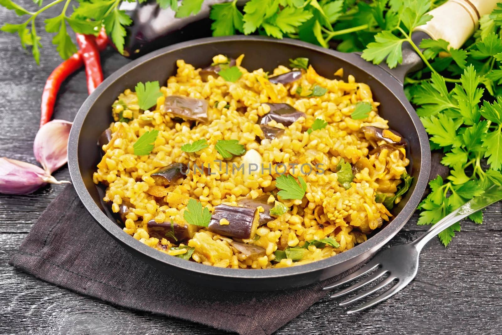 Bulgur stewed with eggplants, carrots, garlic and onions in a frying pan on a towel against the background of dark wooden board