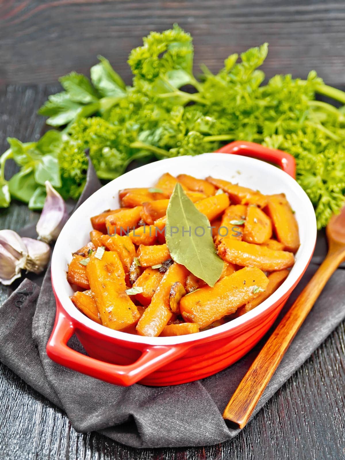 Carrots fried with garlic, bay leaf, spices and green onions in a pan on a towel, basil and parsley on dark wooden board background