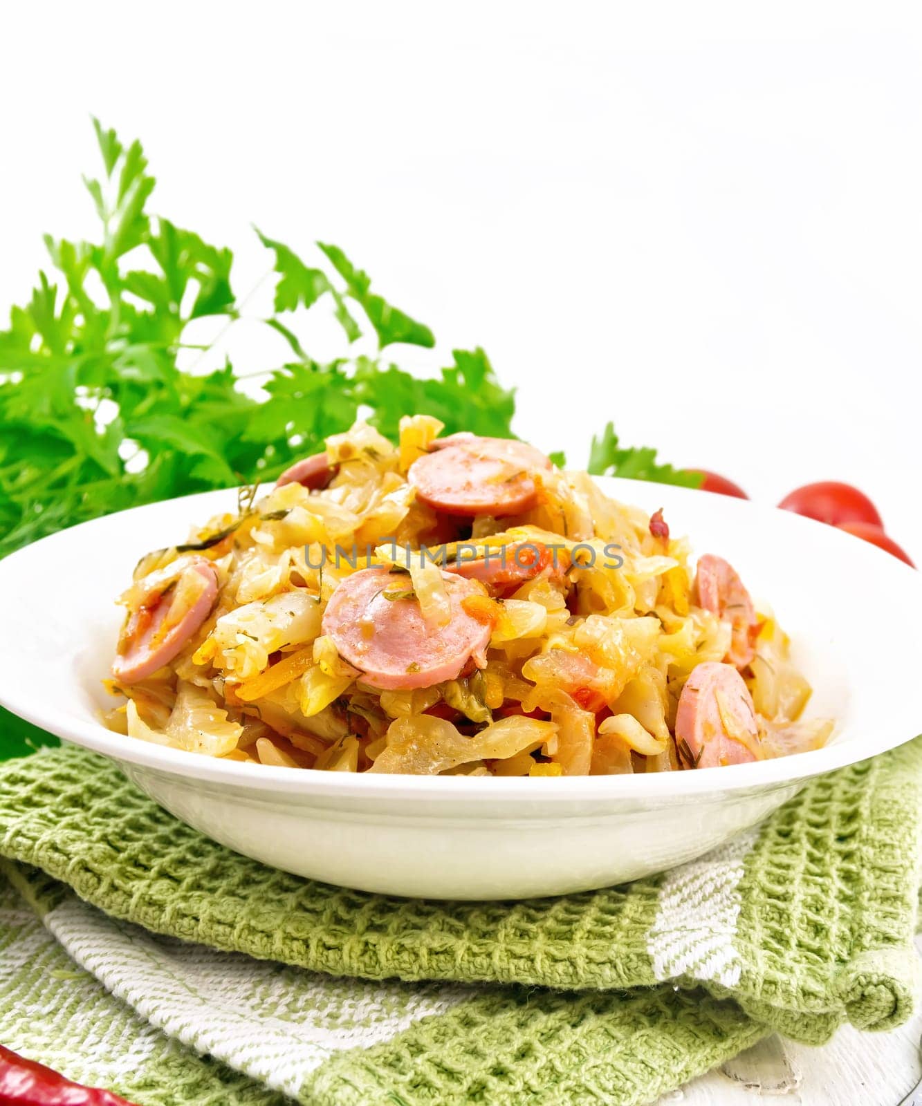 Cabbage stew with sausages in a white plate on a towel, tomatoes, parsley and a fork on the background of light wooden boards