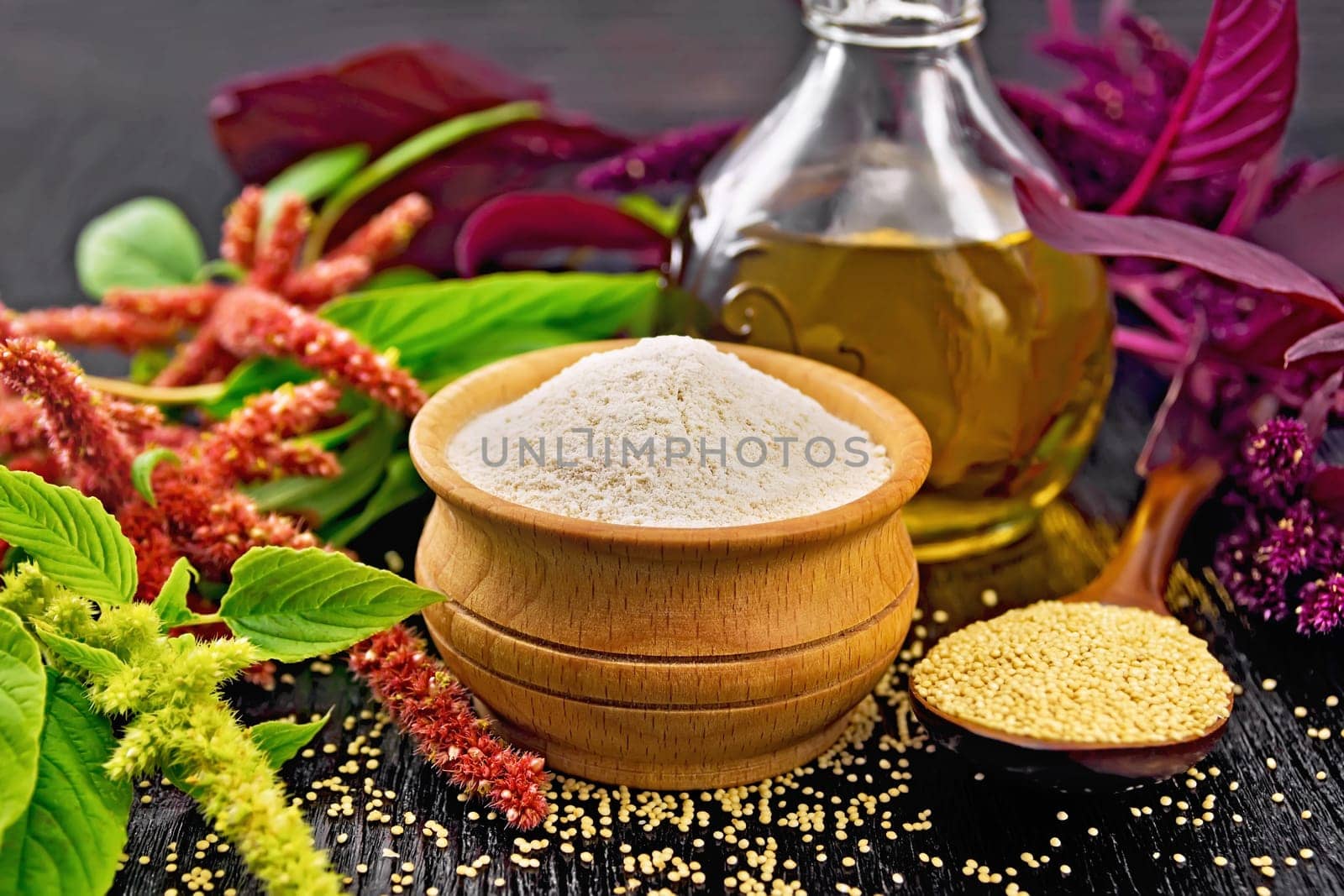 Amaranth flour in a bowl, seeds in a spoon and oil in decanter, brown, green and purple flowers of plant on black wooden board background