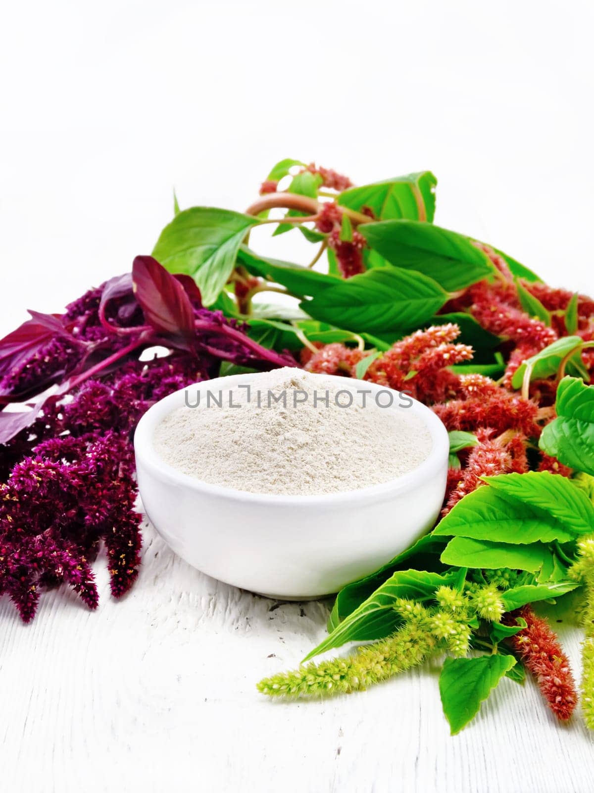 Amaranth flour in a bowl, brown, green and purple flowers of a plant on light wooden board background