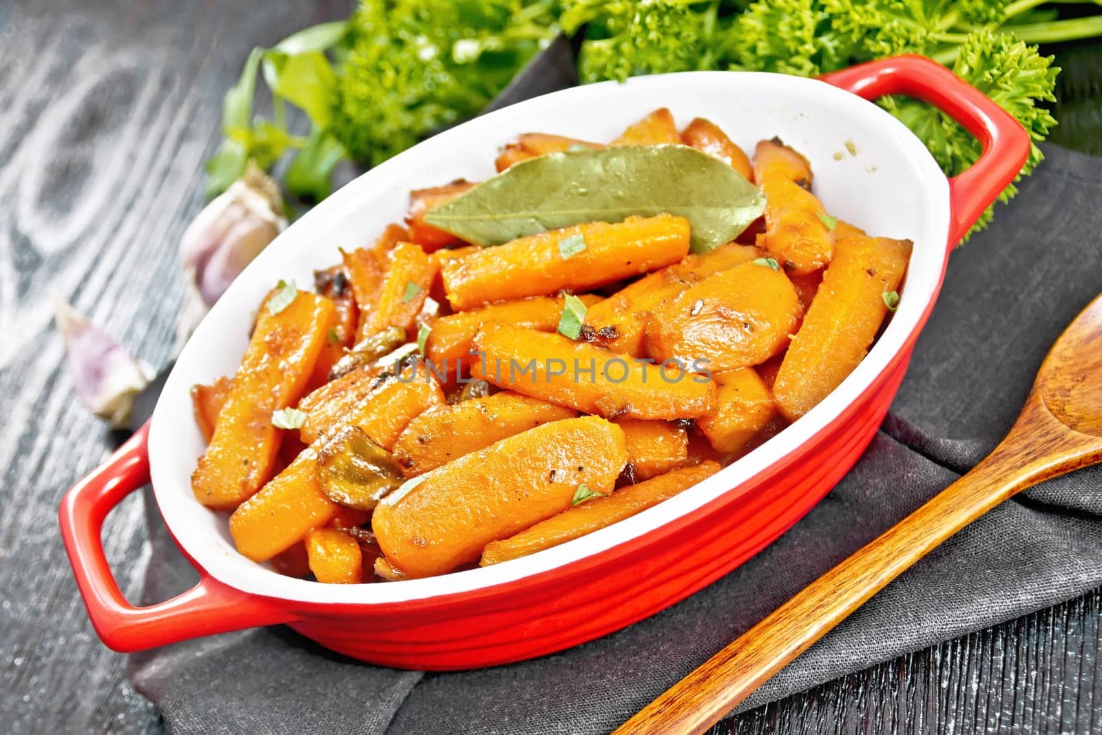 Carrots fried with garlic, bay leaf, spices and green onions in a pan on a napkin, basil and parsley on black wooden board background