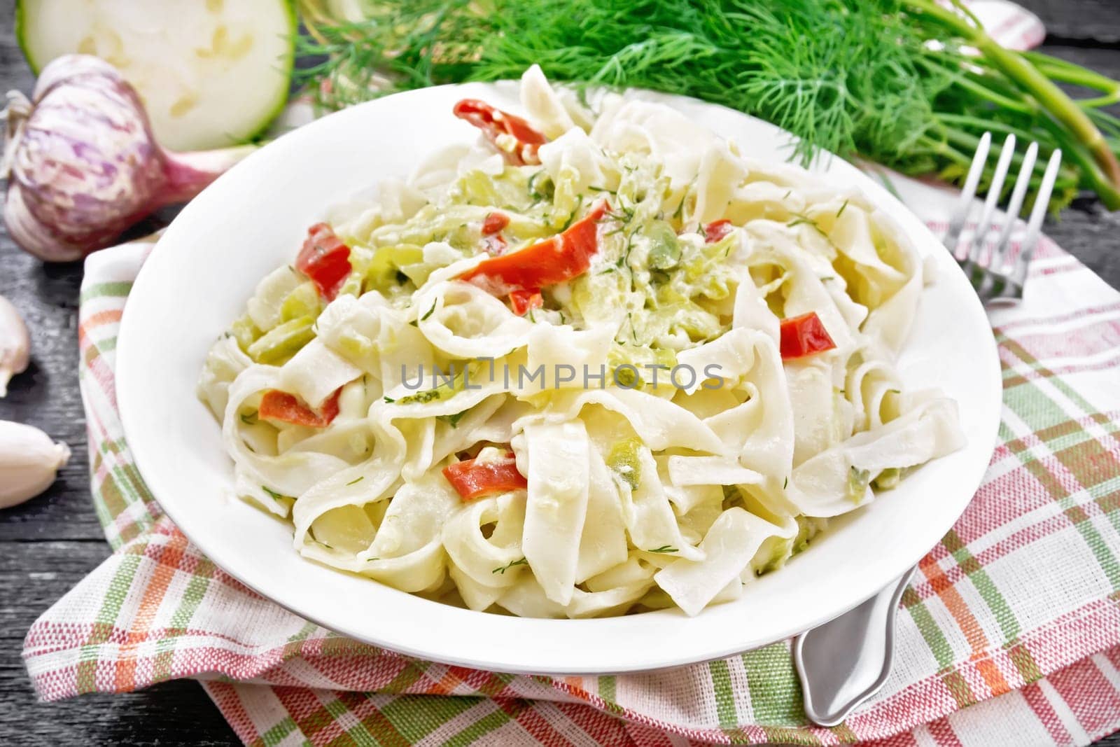 Fettuccine pasta with zucchini and hot red pepper in creamy sauce in white plate on a towel, garlic and a fork on black wooden board background