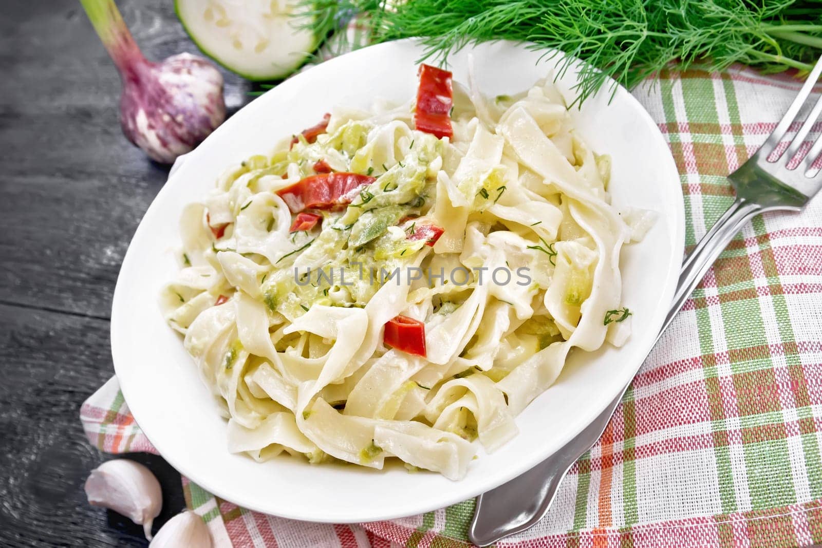 Fettuccine pasta with zucchini and hot red pepper in creamy sauce in white plate on a towel, garlic and a fork on wooden board background