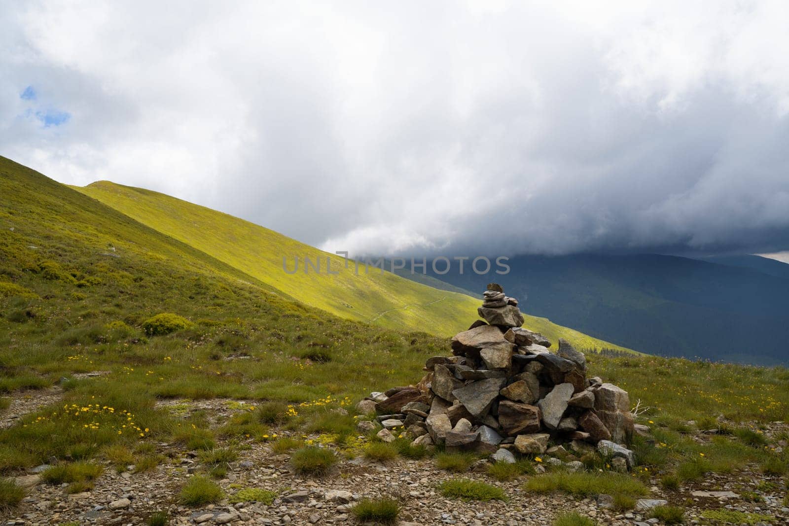 closeup of pyramid of stacked stones used for hiking trail tourist guidance with beautiful sunny mountain slope range covered in green grass in the background and cloudy sky