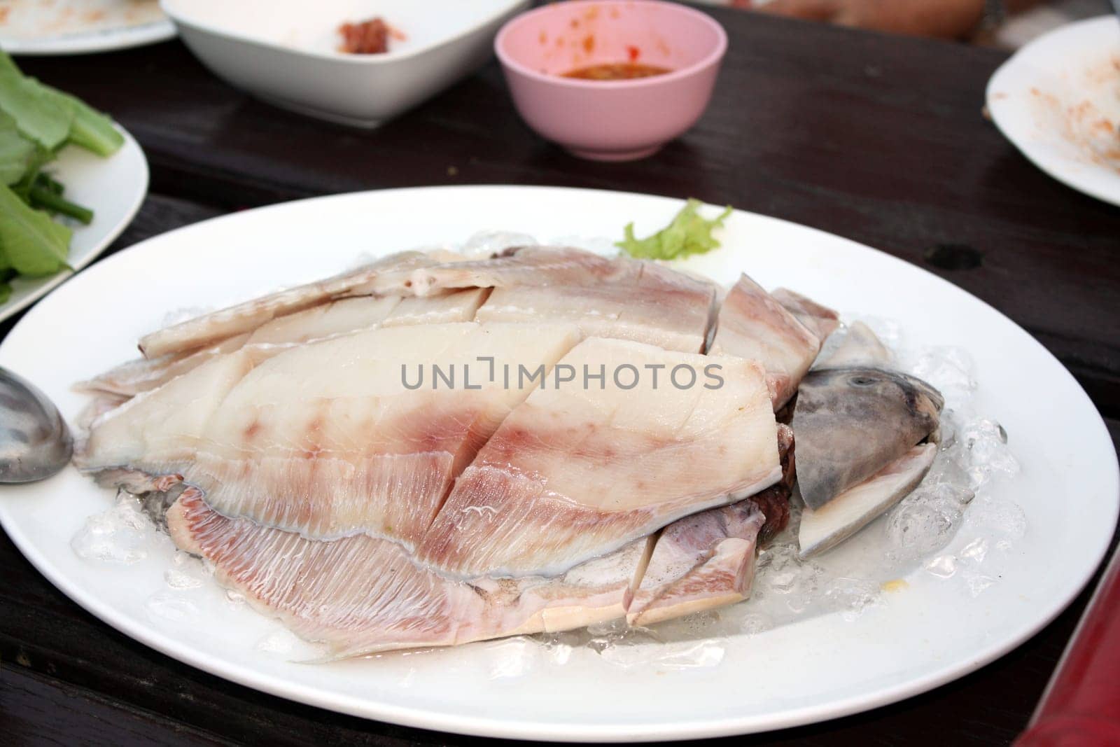 Fresh Gray pomfret, Chinese silver pomfret fish cut into pieces arranged in a white plate topped with ice. On the wooden table in the restaurant, prepare for the shabu pot. Selective focus.