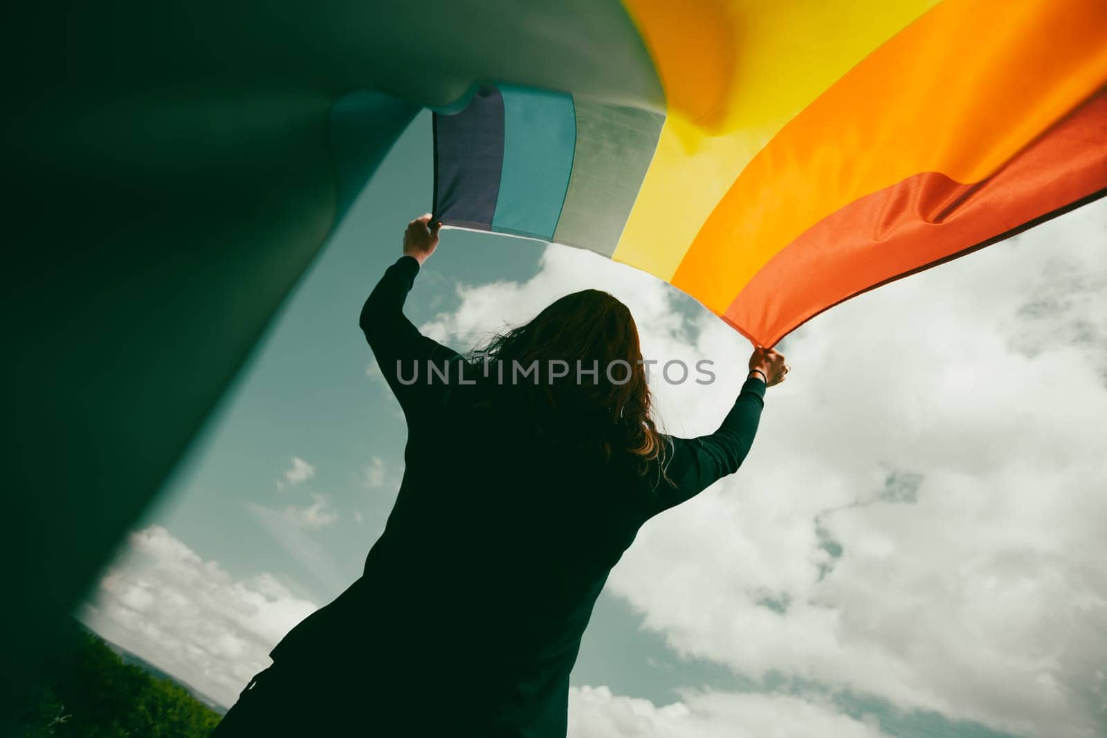 Woman sitting in a green meadow holding a Gay Rainbow Flag. Bisexual,gay, lesbian, transsexual symbol. Happiness, freedom and love concept for same sex couples