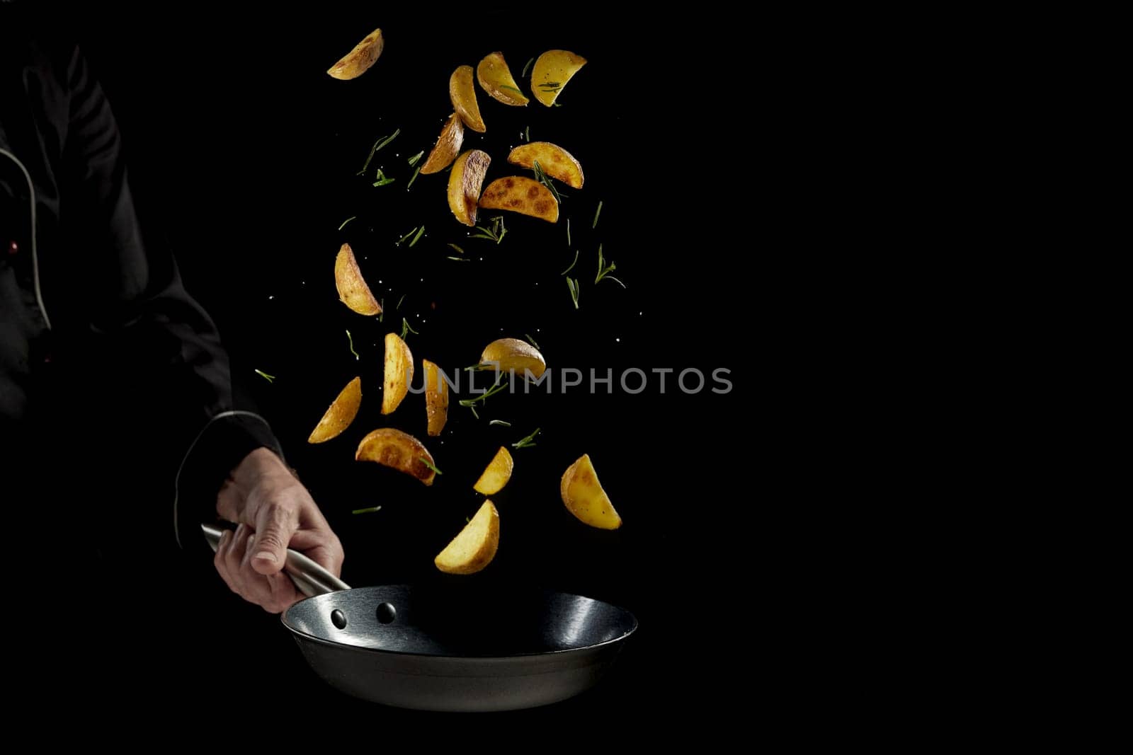 Crop faceless male chef in uniform tossing delicious cut potatoes and herbs in frying pan while preparing appetizing dinner against black background