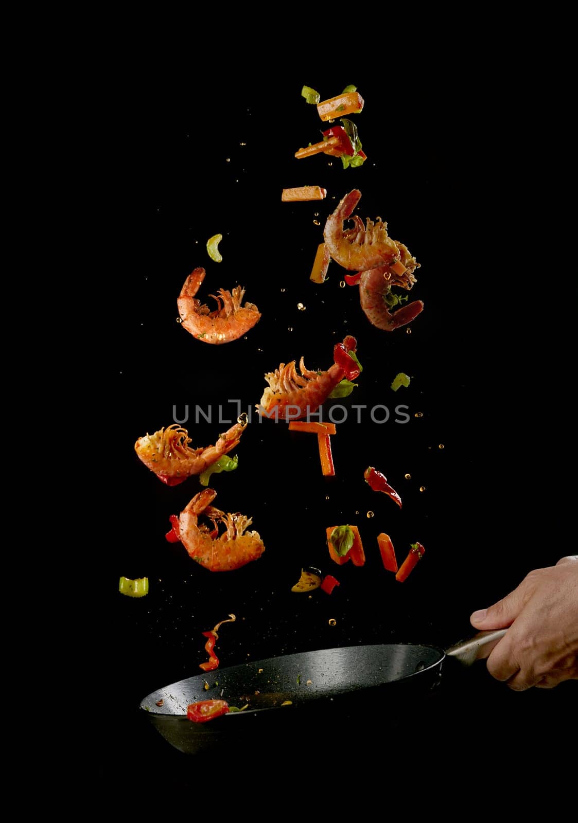 Crop anonymous male chef tossing food in frying pan while preparing delicious prawns and vegetables against black background