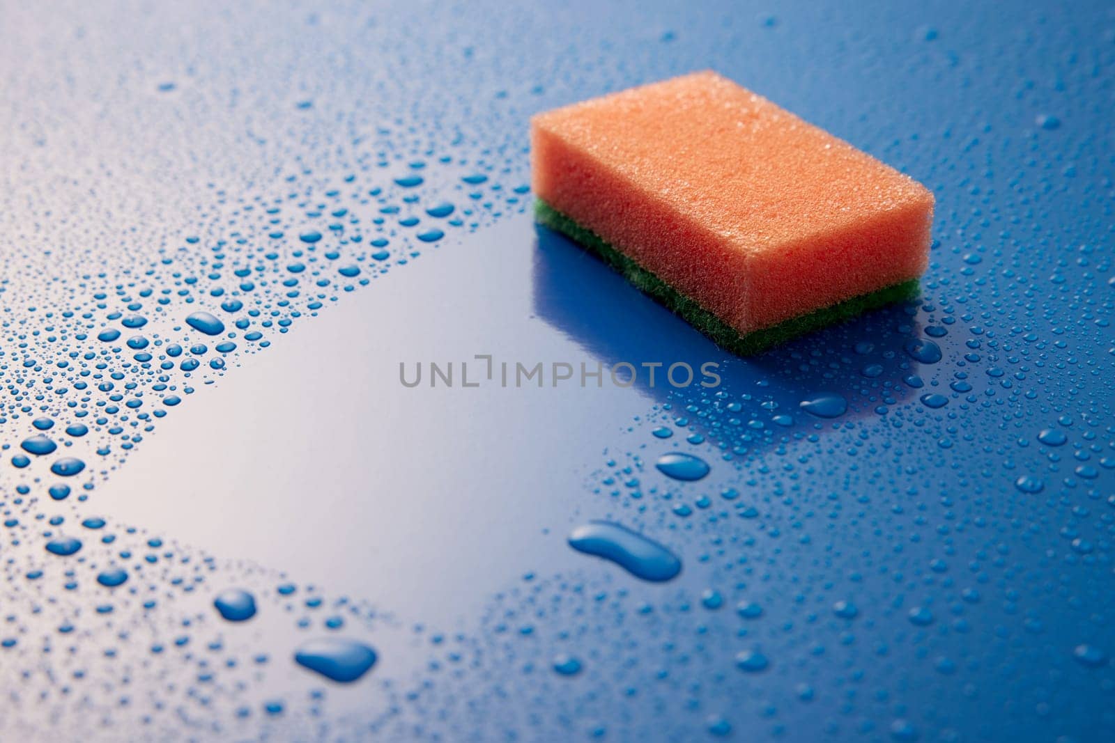 Orange scrub sponge placed on wet blue surface covered with small drops of water in light room during daily hygiene routine
