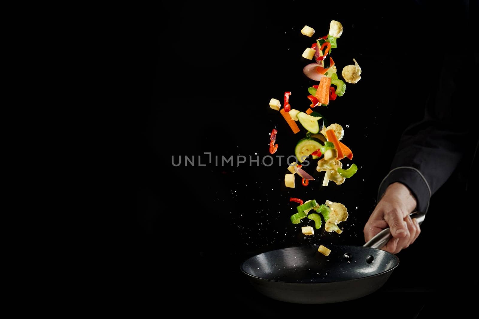 Crop unrecognizable male chef in uniform throwing fresh delicious assorted vegetables while preparing healthy dinner in frying pan against black background