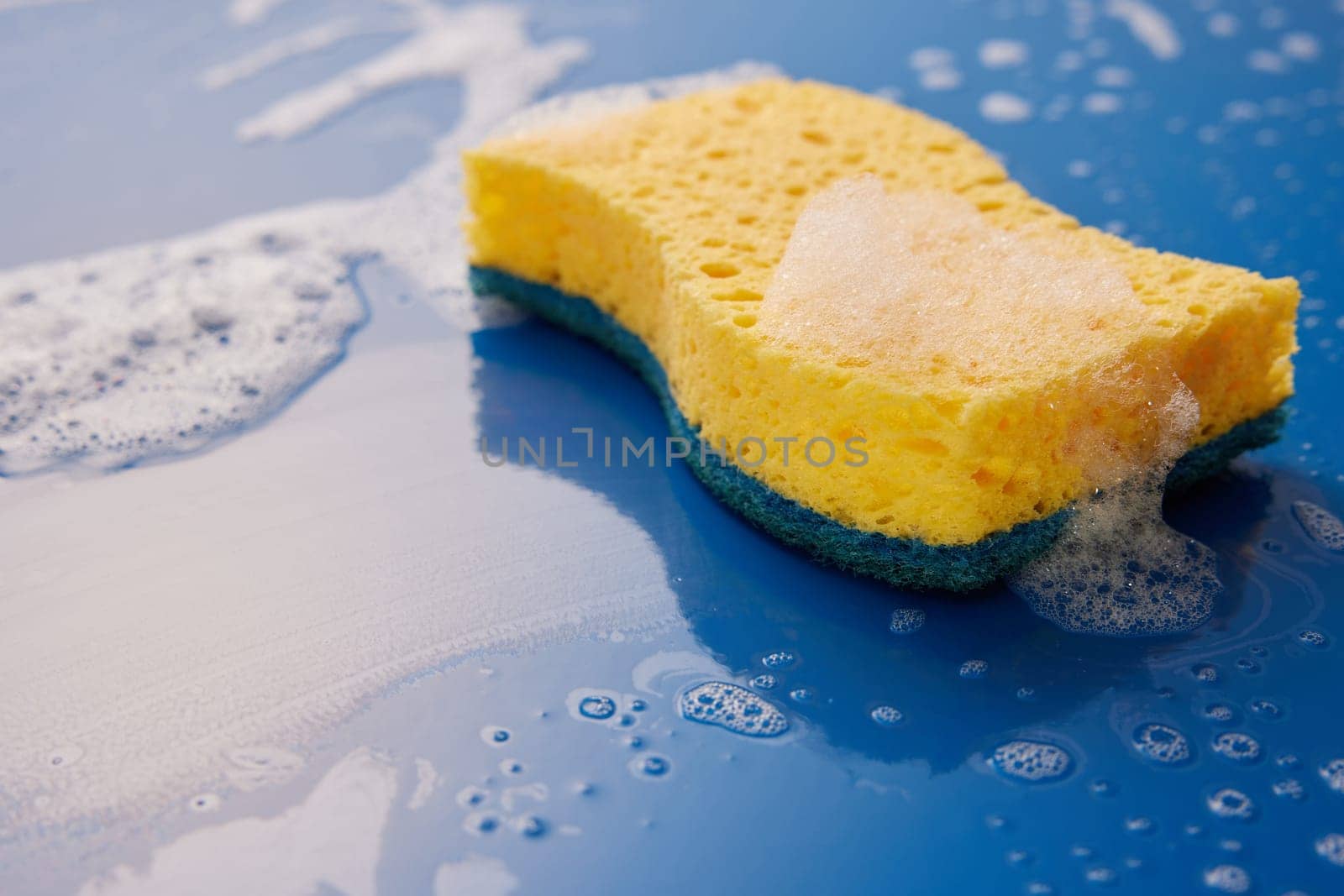 Yellow wash scrub sponge for dishwashing placed on wet blue background with soap bubbles in light room during household chores