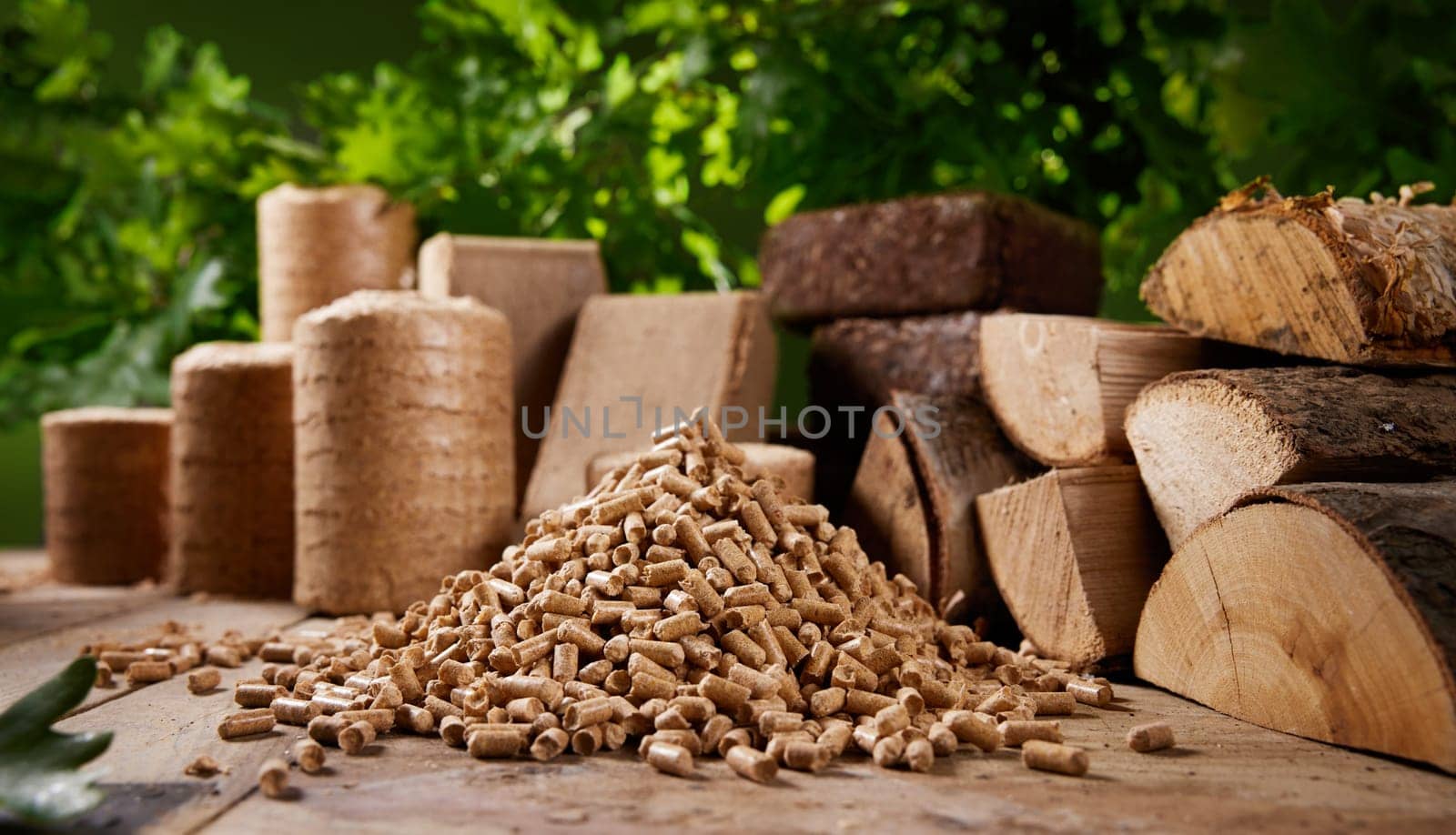 Pyramid of biofuel pellets stacked on wooden table near biomass briquettes and chopped firewood against lush green oak tree