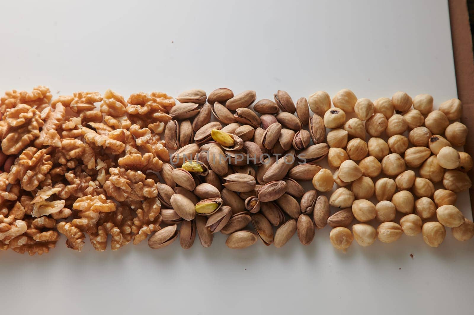 From above pile of walnuts and pistachios placed near hazelnuts arranged in neat rows on white table in light room