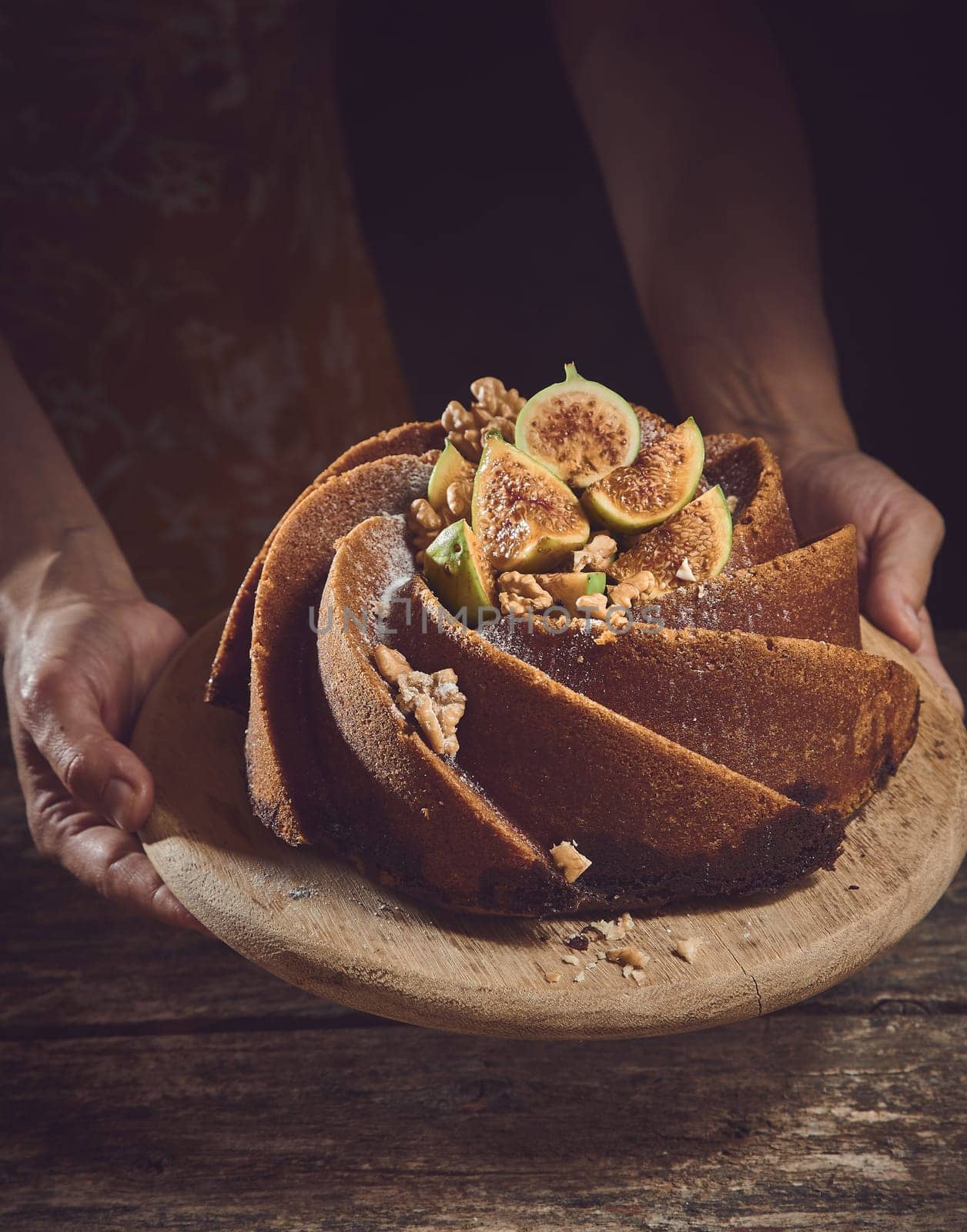 Crop unrecognizable cook holding sweet freshly baked cake made of figs with decorations on wooden cutting board in light kitchen