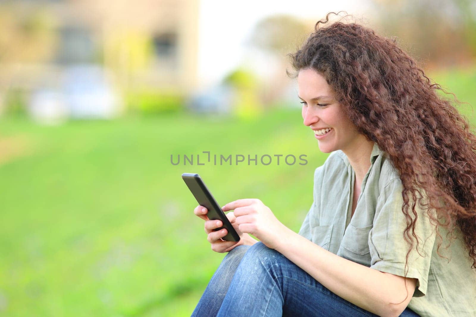Happy woman with curly hair sitting using smart phone on the grass in a park