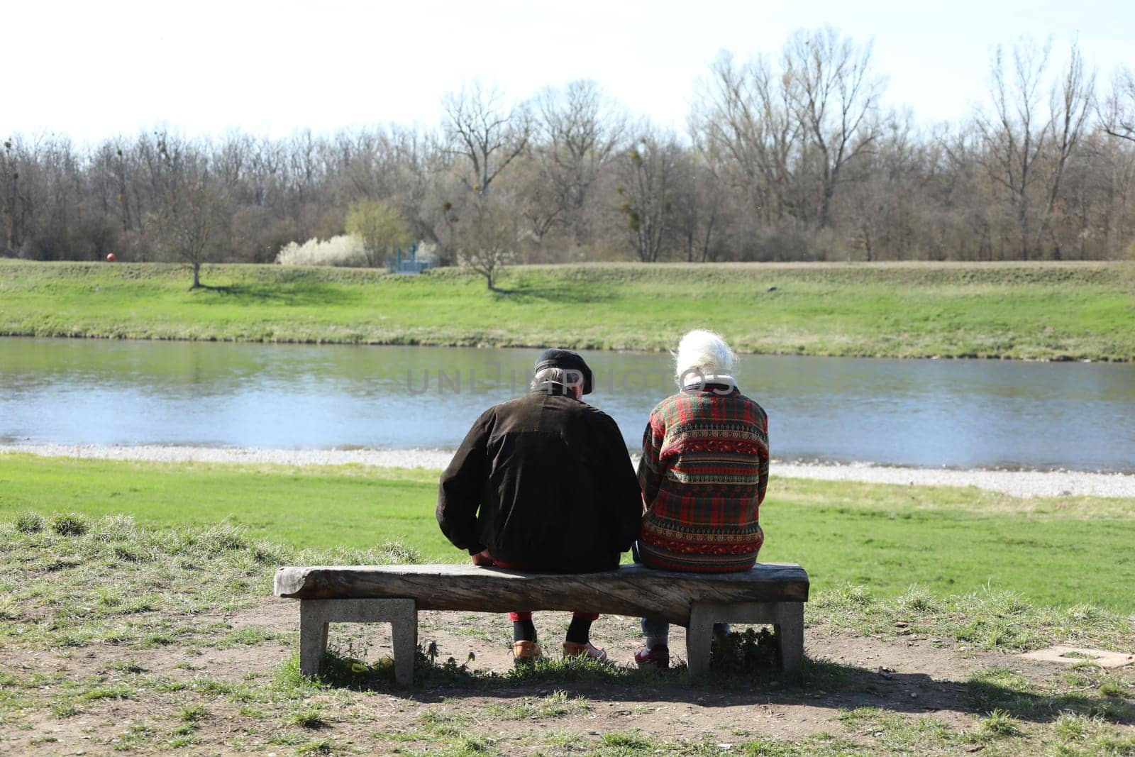 Two seniors enjoy the warming spring sun on a wooden bench on the banks of the Rhine near Hartheim.