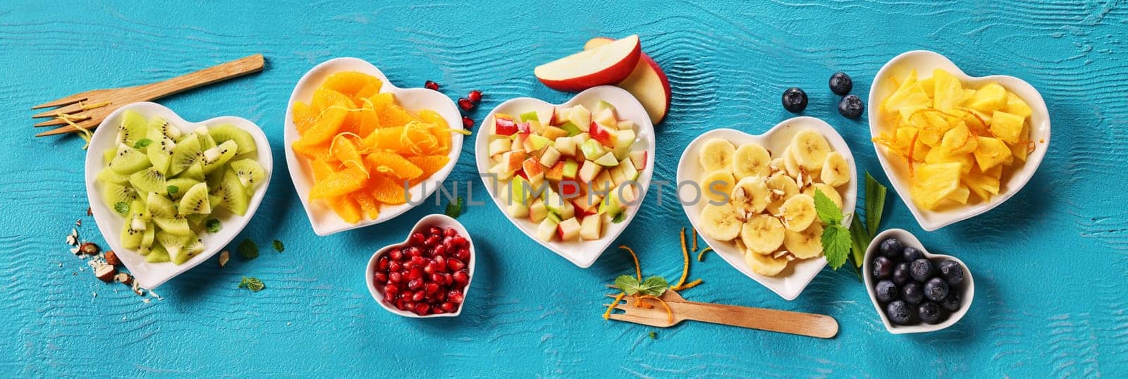 Top view of mix of fresh appetizing chopped fruits and berries served in heart shaped bowls on blue background with wooden forks