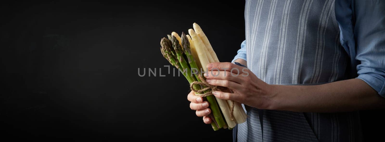 Crop anonymous female cook with uncooked healthy green and white asparagus standing on black background in studio during cooking preparation