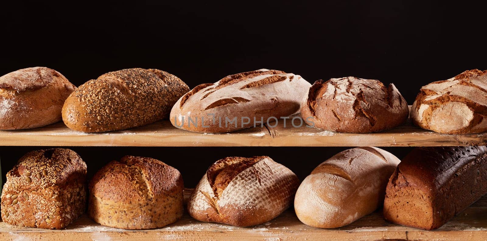 Bunch of appetizing freshly baked loaves of bread placed on wooden shelves with flour against black background in light studio