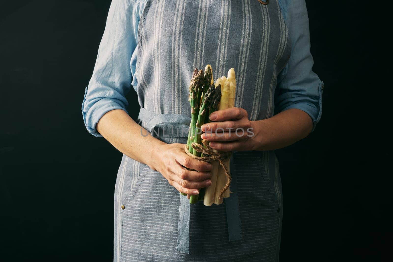 Crop faceless female cook in apron holding mix of raw healthy asparagus against black background during cooking preparation in studio